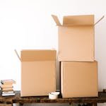 Opened cardboard boxes and stack of books placed on wooden shabby table near tape with scissors against white plain wall